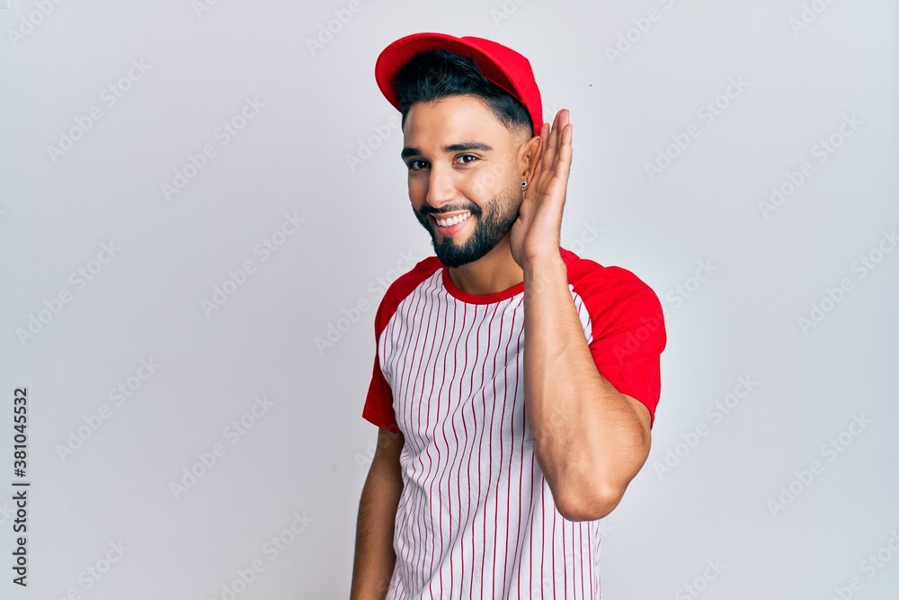 Young man with beard wearing baseball uniform smiling with hand over ear listening and hearing to rumor or gossip. deafness concept. Young man with beard wearing baseball uniform smiling with hand over ear listening and hearing to rumor or gossip. deafness concept.