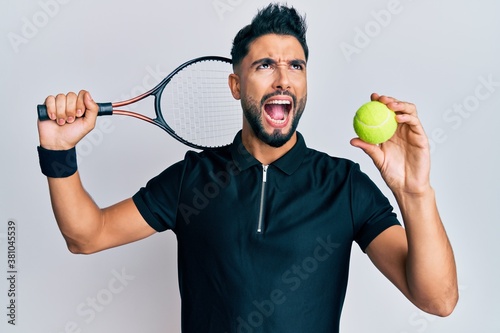 Young man with beard playing tennis holding racket and ball angry and mad screaming frustrated and furious, shouting with anger looking up. © Krakenimages.com