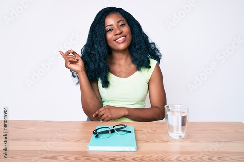 Beautiful african woman sitting on the table stuying for university smiling happy pointing with hand and finger to the side photo