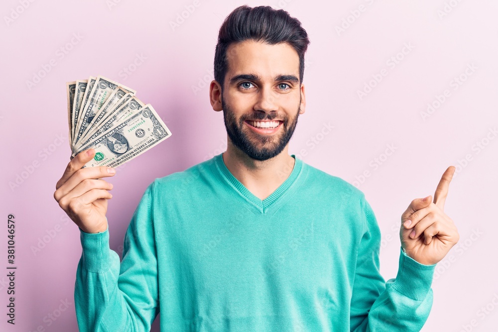 Young handsome man with beard holding bunch of dollar smiling happy pointing with hand and finger to the side Young handsome man with beard holding bunch of dollar smiling happy pointing with hand and finger to the side