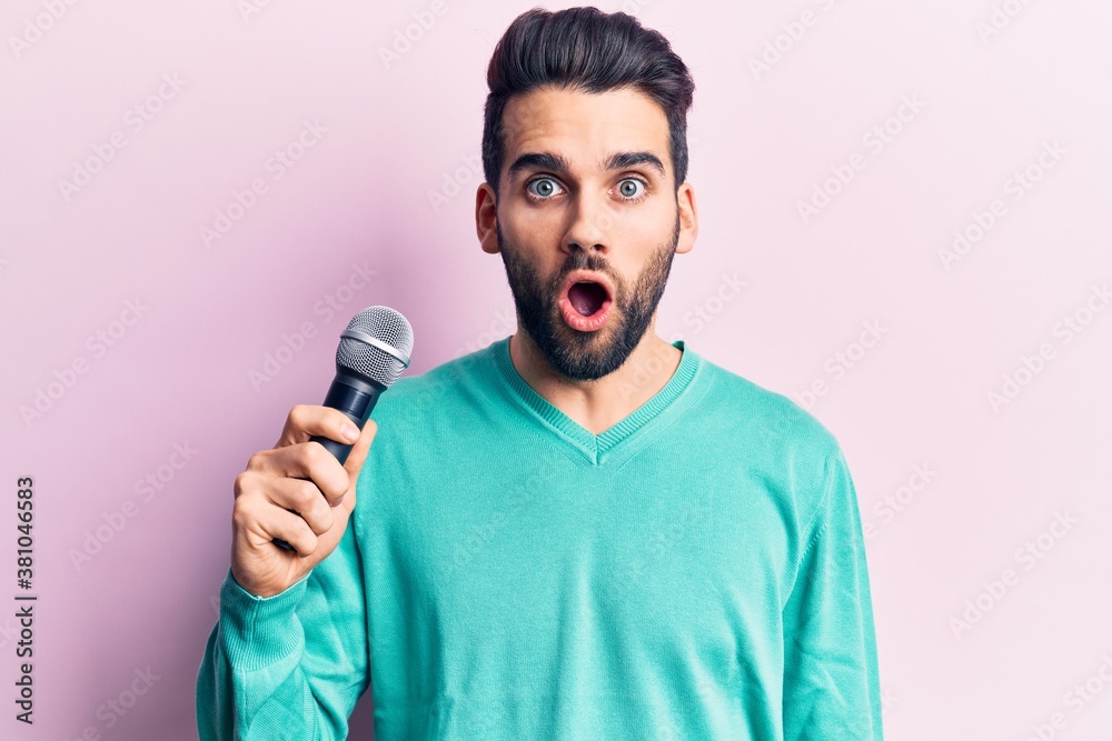 Young handsome man with beard singing song using microphone scared and amazed with open mouth for surprise, disbelief face Young handsome man with beard singing song using microphone scared and amazed with open mouth for surprise, disbelief face