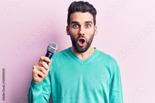 Young handsome man with beard singing song using microphone scared and amazed with open mouth for surprise, disbelief face