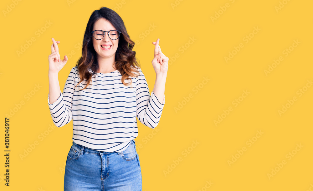 Beautiful young brunette woman wearing casual clothes and glasses gesturing finger crossed smiling with hope and eyes closed. luck and superstitious concept. Beautiful young brunette woman wearing casual clothes and glasses gesturing finger crossed smiling with hope and eyes closed. luck and superstitious concept.