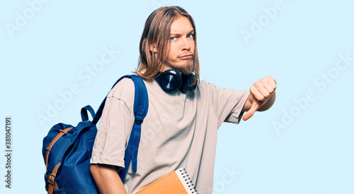 Handsome caucasian man with long hair wearing student backpack and holding books with angry face, negative sign showing dislike with thumbs down, rejection concept