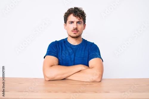 Young caucasian man with curly hair wearing casual clothes sitting on the table skeptic and nervous, disapproving expression on face with crossed arms. negative person.