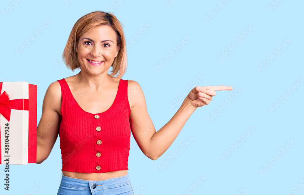 Young blonde woman holding gift smiling happy pointing with hand and finger to the side Young blonde woman holding gift smiling happy pointing with hand and finger to the side