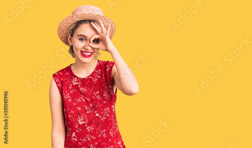 Young beautiful blonde woman wearing summer hat doing ok gesture with hand smiling, eye looking through fingers with happy face.