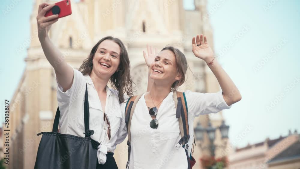Handheld shot of two young cheerful women sitting on a monument in a city center and taking a selfie with a smartphone. Handheld shot of two young cheerful women sitting on a monument in a city center and taking a selfie with a smartphone.