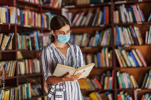 Young attractive female student in dress with brown hair standing in the library with mask on face and reading a book. Studying during corona virus pandemic.