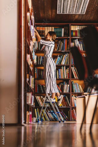 Konstfotografi Full length of young attractive librarian standing on ladder and searching for a book