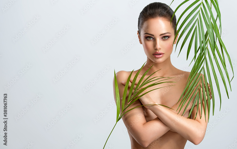 Young beautiful woman with healthy skin of body and palm leaves. Tanned body of an attractive white girl with green plants. Woman hiding her naked body with hands. Young beautiful woman with healthy skin of body and palm leaves. Tanned body of an attractive white girl with green plants. Woman hiding her naked body with hands.