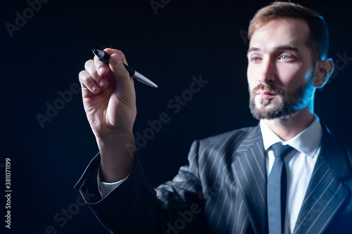 Man writes something on screen. Businessman raised his hand to screen. Concept - businessman writes on a virtual screen. Businessman held out a pen to camera. Guy in a dell suit on black background.