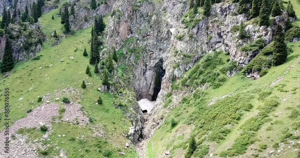 Black waterfall in a gorge with an ice cave. Top view of green hills and huge rocks. Remnants of an avalanche in the gorge. Fir trees and bushes grow on the rocks. Huge snow cave. Kazakhstan, Tuyuk Su Black waterfall in a gorge with an ice cave. Top view of green hills and huge rocks. Remnants of an avalanche in the gorge. Fir trees and bushes grow on the rocks. Huge snow cave. Kazakhstan, Tuyuk Su