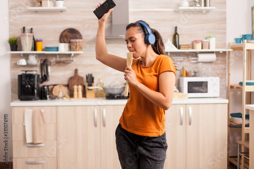 Young beautiful woman dancing while listening music in blue wirelless headphones in the kitchen. Energetic, positive, happy, funny and cute housewife dancing alone in the house. Entertainment and