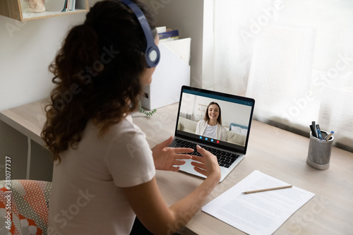 Top back view of female student in headphones sit at desk at home talk on video call with teacher or trainer. Woman in earphones have webcam conference, study distant on computer. Education concept.
