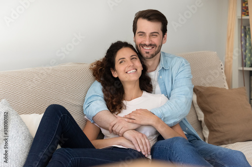 Overjoyed young Caucasian couple tenants relax on couch at home hugging look in distance dreaming visualizing. Happy renters rest on sofa embrace, enjoy family weekend in new house. Realty concept.