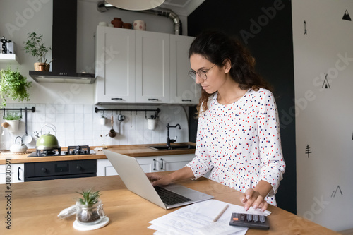 Concentrated young Caucasian wife look at laptop screen manage household expenses expenditures on computer. Focused millennial woman pay bills taxes on laptop online, calculate family budget at home.