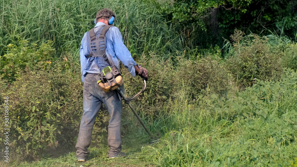 Petrol brush cutter. Man in overalls, protective glasses, soundproof headphones and work gloves mows the grass with gas cutter. Full-length rear view.
