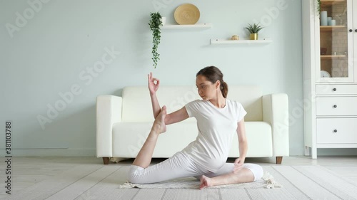 Calm young pregnant brunette woman does ardha matsyendrasana practicing yoga position on floor near sofa in spacious room at home photo