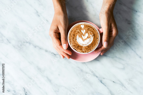 Pink cup of fresh cappuccino in woman hands on white marble table background. Classic latte art and chocolate on foam. Empty place for text, copy space. Coffee addiction.