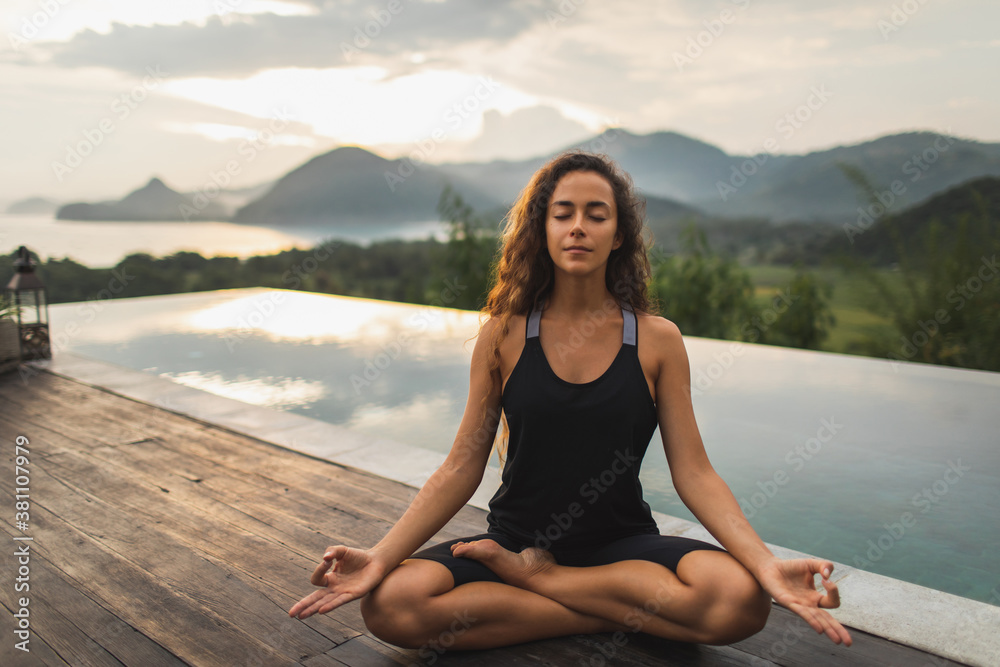 Woman practicing yoga in lotus position on infinity poolside with beautiful ocean and mountain view in morning. Healthy lifestyle, spiritual and emotional concept. Awakening.