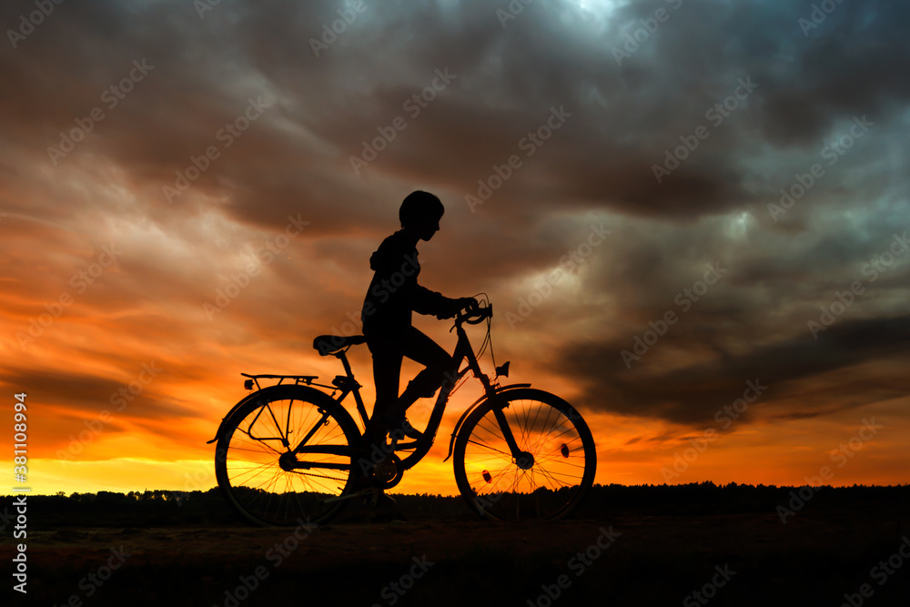 Fototapeta premium Boy , kid 10 years old, riding bike in countryside in dramatic cloudy sky background, silhouette of riding person at sunset in nature