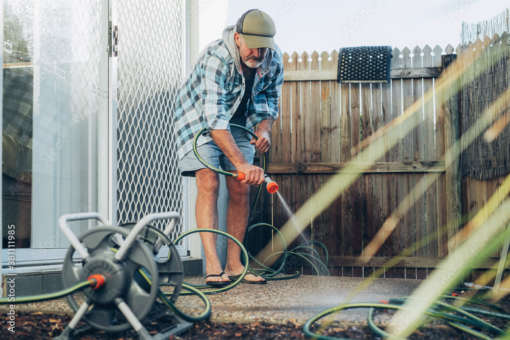 Caucasian adult middle aged bearded man cleans outdoor terrace with flow of water from hose pipe