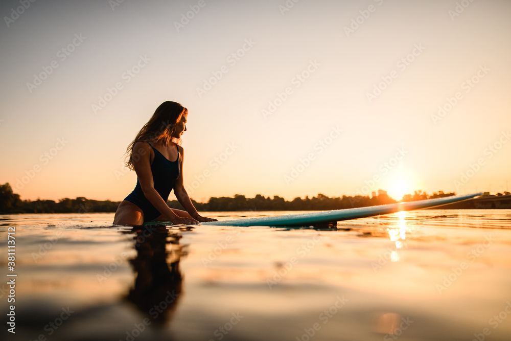 beautiful woman sitting on surf style wakeboard on water against background of sunrise
