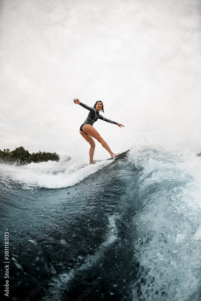 view on young sportive woman confidently stands on the wake surf board and rides the wave.