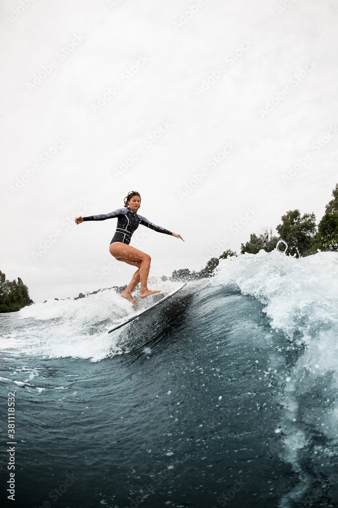 athletic woman in black wetsuit effectively jumps on surf board on wave