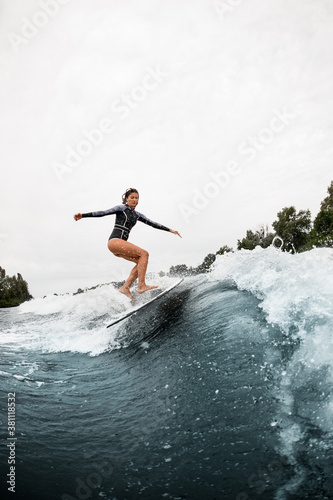 athletic woman in black wetsuit effectively jumps on surf board on wave