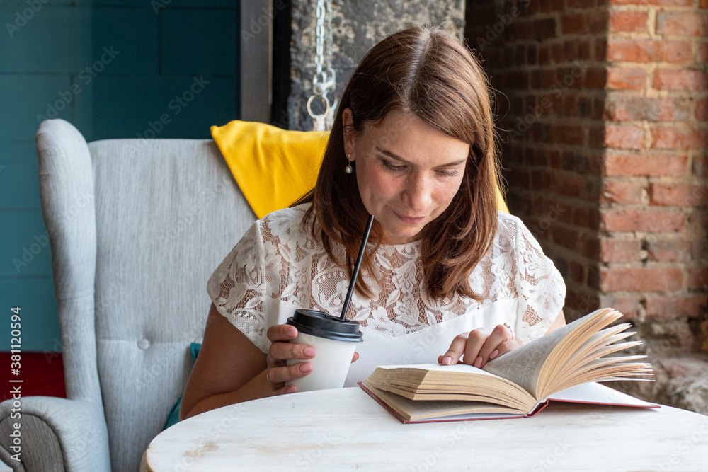 Obraz premium A beautiful young woman with red long hair in a white blouse sits in a chair in cafe , drinks coffee and reads a book