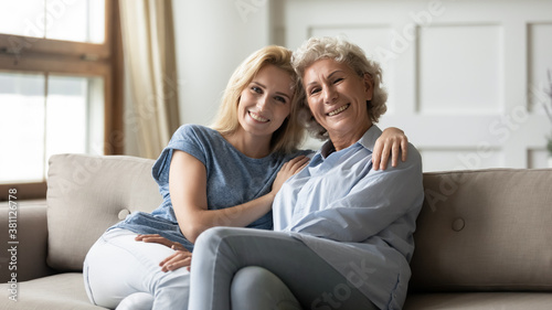 You are like family to me. Portrait of happy senior grey-haired lady sitting on sofa in living room hugging warm young female au pair or home care assistant trusting her, smiling and looking at camera
