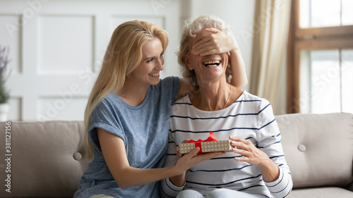 Guess what. Laughing grown up daughter congratulates happy mature mother covering her eyes and giving birthday gift, excited senior grandma receiving surprise on Mothers Day from adult granddaughter