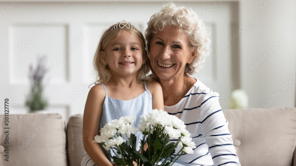 Happy smiling old grandmother posing for portrait on Mothers Day looking at camera with sweet little grandkid girl on laps, embracing her with affection and holding bunch of tender spring flowers Happy smiling old grandmother posing for portrait on Mothers Day looking at camera with sweet little grandkid girl on laps, embracing her with affection and holding bunch of tender spring flowers