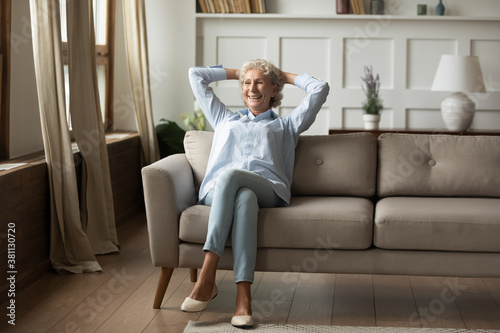 Home, sweet home. Happy smiling retired senior female relaxing at living room indoor sitting alone cross-legged on cozy sofa with hands behind head imagining or remembering pleasant things, copy space