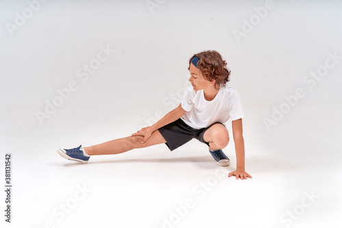 Being physically active. Full length of caucasian teenage boy in sportswear stretching legs, warming up before training isolated over grey background in studio