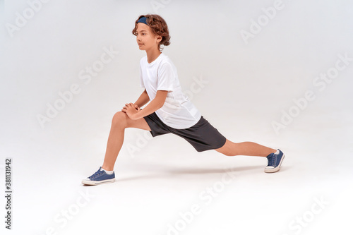 Stretching exercises. Full length of caucasian sporty teenage boy in sportswear warming up before training, standing isolated over grey background in studio