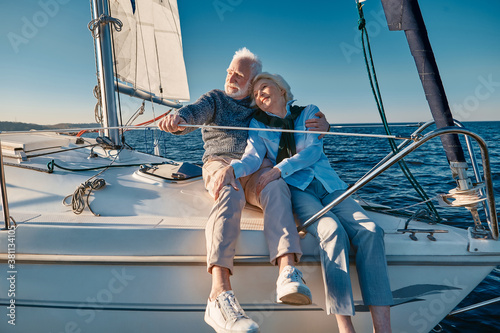 Enjoying sailing. Happy lovely senior family couple hugging and relaxing on a sail boat or yacht deck floating in a calm blue sea, looking at the horizon