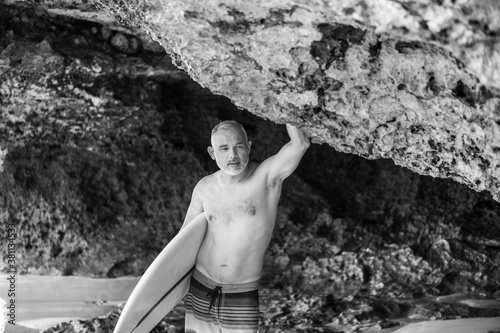 Black and white portrait of handsome shirtless man surfer   holding white surf board  and cliff  rocks on background