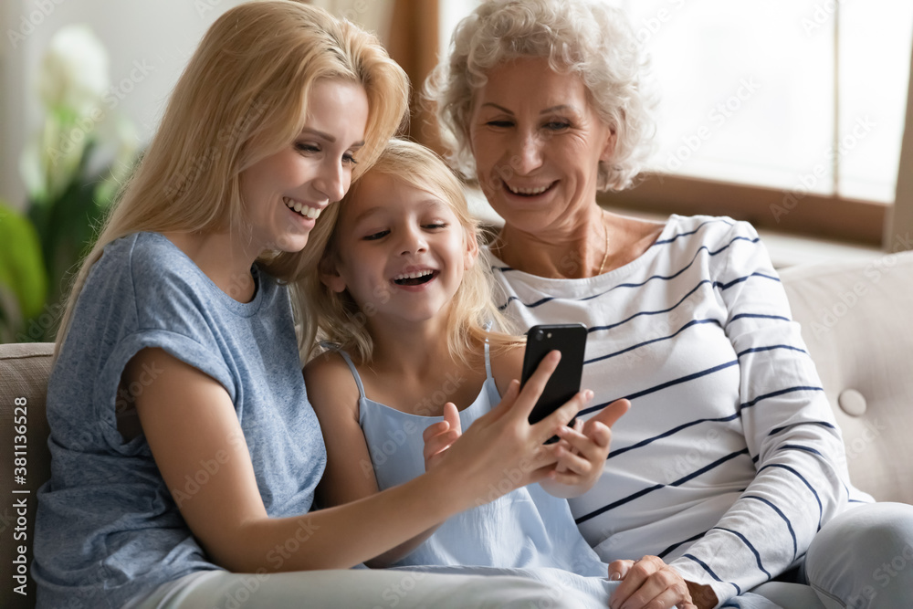 Cute thing. Happy laughing little kid girl relaxing on couch on weekend with senior grandmother and adult mom reading internet blog on cell, playing online game, sharing photos, enjoying video by wifi Cute thing. Happy laughing little kid girl relaxing on couch on weekend with senior grandmother and adult mom reading internet blog on cell, playing online game, sharing photos, enjoying video by wifi