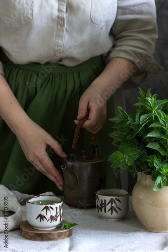 Mint tea in a vintage Chinese tea set, a woman in a linen dress and an apron, and a bouquet of mint in a jug.