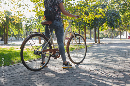 A girl in jeans and sneakers stands next to a beige bicycle in a park among green trees and a lawn