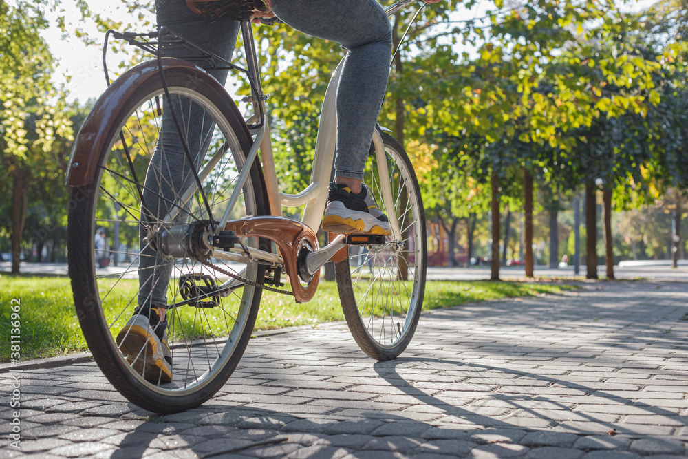 A girl in jeans and sneakers sits on a beige bicycle in the park among green trees and lawn A girl in jeans and sneakers sits on a beige bicycle in the park among green trees and lawn