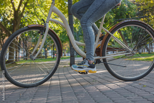 A girl in jeans and sneakers rides a beige bike in the park among green trees and lawn