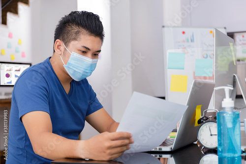 Asian small business start up working at Private Home Office social distancing among Coronavirus outbreak. Businessman wear face mask holding document paper chart .