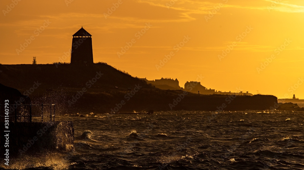 A glorious orange sky in the morning silhouettes the remains of a coastal windmill as the waves crash into the shore