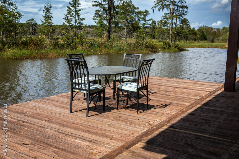 A set of black metal chairs and a table sitting on a large wooden dock deck outside of a hunting and fishing cabin near a large pond in Georgia for outdoor dining
