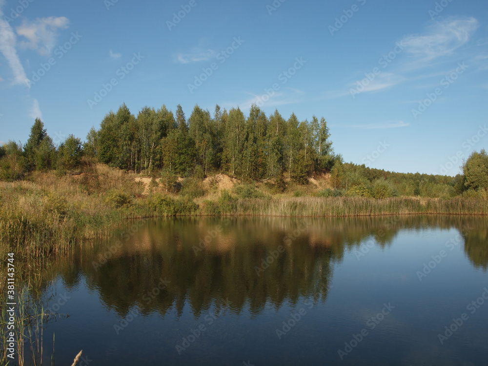Autumn landscape. The sandy coast overgrown with forest is reflected in the water against the background of the autumn sky.