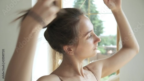 sporty brunette pretty female stands at window, fixes her hair, collects hair in bun. closeup shot of young caucasian woman standing in living room photo